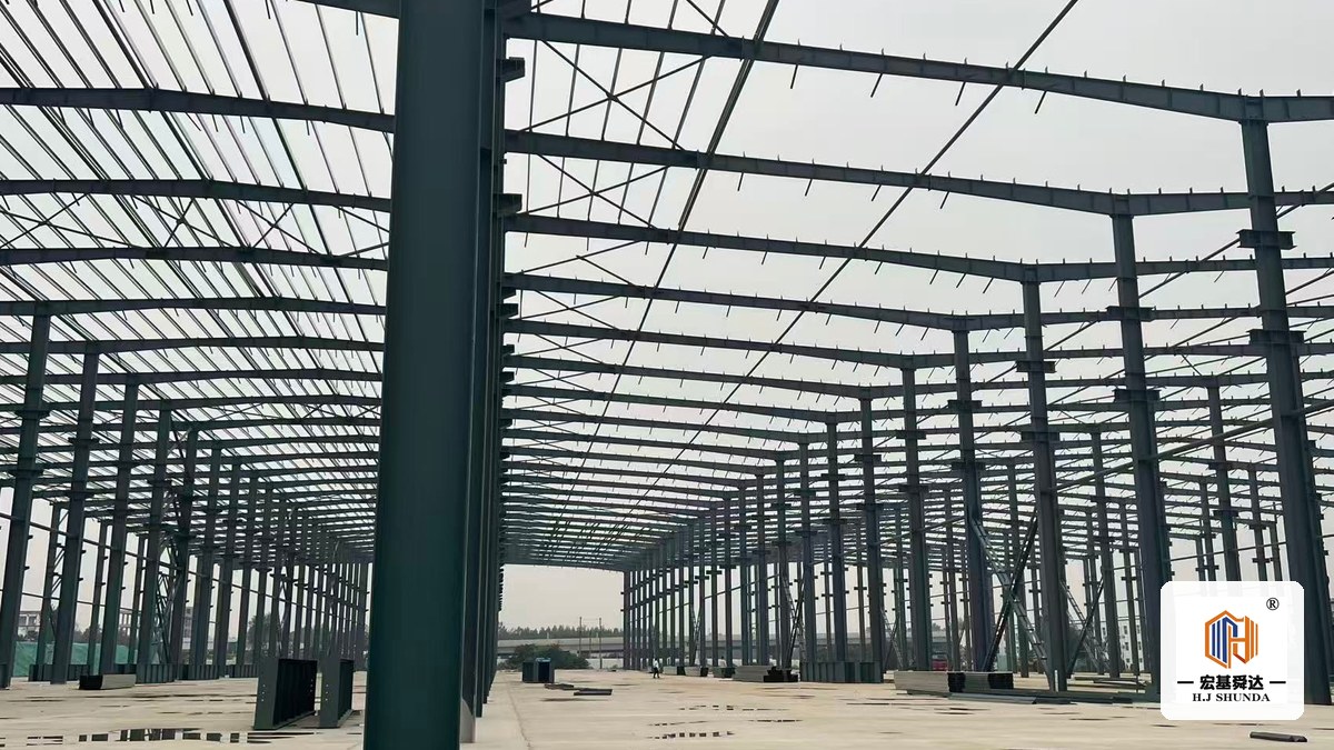 Erected steel portal frames running the length of a large workshop, with a main column standing in the foreground.